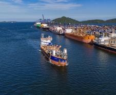 #ParaQuemNaoPodeVer A imagem mostra uma fotografia de um porto com vários barcos atracados. A água do mar é de um azul intenso e há uma grande quantidade de barcos coloridos, principalmente em tons de branco e azul, ao longo de um cais. Ao fundo, é possível ver uma linha de montanhas sob um céu azul claro.