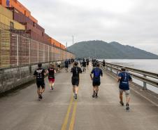 #praquemnaopodever vários corredores aparecem de costas cruzando o cais, pelas margens do Porto de Paranaguá. À direita, aparecem as aguas da baía de Paranaguá, ao fundo as ilhas cobertas de árvores e à esquerda os contêineres amarelos, vermelhos e laranjas.