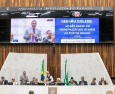 #praquemnaopodever 10 pessoas estão sentadas em um púlpito alto e com fachada branca. Em pé está um homem de terno cinza e gravata rosa, segurando um microfone na mão e discursando enquanto gesticula com a mão direita. Seu perfil aparece no telão ao fundo, ao lado de um texto com os dizeres "sessão solene em homenagem aos 90 anos do Portos Paraná"