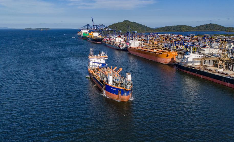#ParaQuemNaoPodeVer A imagem mostra uma fotografia de um porto com vários barcos atracados. A água do mar é de um azul intenso e há uma grande quantidade de barcos coloridos, principalmente em tons de branco e azul, ao longo de um cais. Ao fundo, é possível ver uma linha de montanhas sob um céu azul claro.